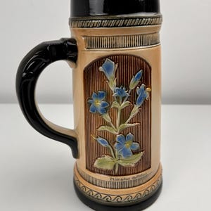 Może przedstawiać: Ceramiczny kufel do piwa z czarnym uchwytem i obramowaniem. Korpus jest w kolorze beżowym z brązowym kwiatowym wzorem i niebieskimi kwiatami. Na podstawie napis "Mimulus guttatus".