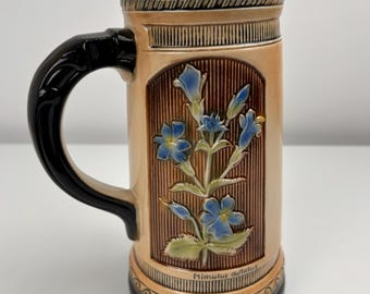 Może przedstawiać: Ceramiczny kufel do piwa z czarnym uchwytem i obramowaniem. Korpus jest w kolorze beżowym z brązowym kwiatowym wzorem i niebieskimi kwiatami. Na podstawie napis "Mimulus guttatus".