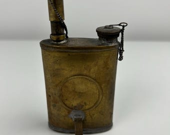 Op de afbeelding: Antieke messing oliekruik met een cilindrisch lichaam, een tuit en twee schroefdoppen. De kruik heeft een aangetaste, verouderde uitstraling, met een kleine ketting bevestigd aan een van de doppen. Het ontwerp omvat een cirkelvormige inkeping aan de voorkant.