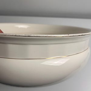 May include: A cream-coloured ceramic bowl with a rounded silhouette and a wide rim. The bowl features a thin gold-coloured band encircling the middle section and a gold-coloured trim along the top edge, which shows signs of wear and fading.