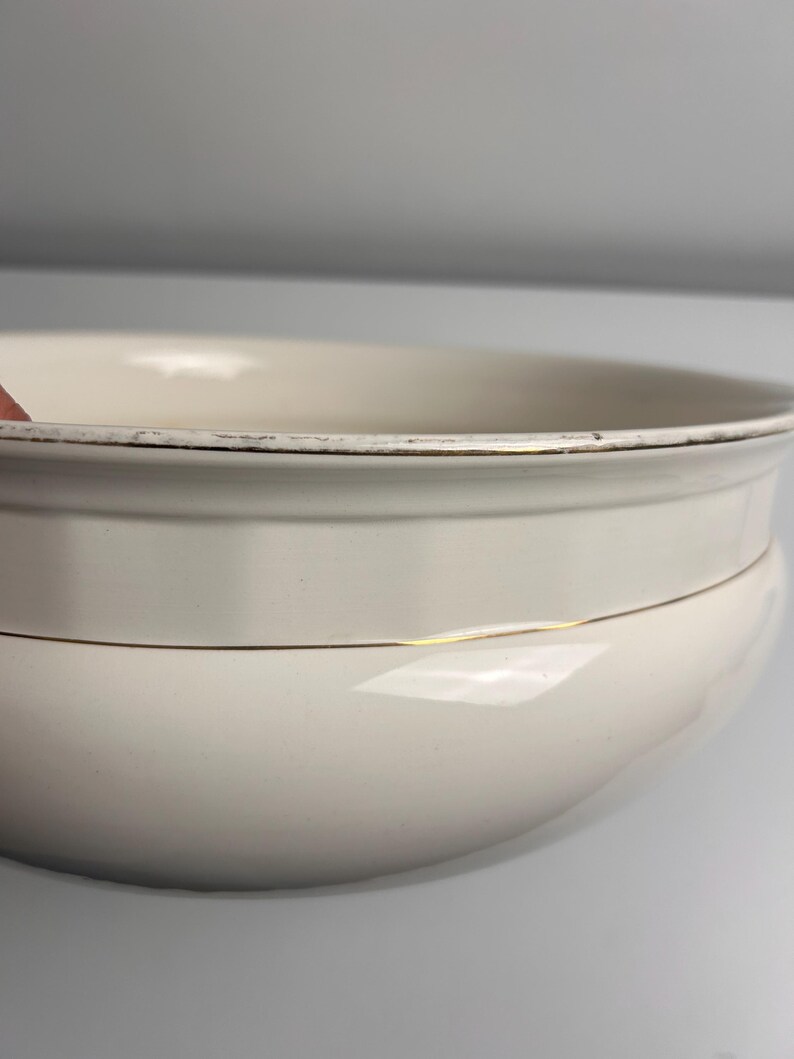 May include: A cream-coloured ceramic bowl with a rounded silhouette and a wide rim. The bowl features a thin gold-coloured band encircling the middle section and a gold-coloured trim along the top edge, which shows signs of wear and fading.