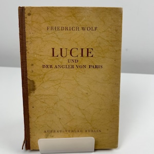 Peut inclure: Un livre vintage intitulé "Lucie und Der Angler von Paris" de Friedrich Wolf. La couverture est de couleur beige délavé avec des lettres rouges. Le livre est maintenu par un support en plastique transparent.