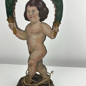 May include: A vintage wooden cherub figurine, hand-painted with flesh tones and brown hair, holding a green wreath with gold accents. The cherub stands on a dark wooden base, wrapped with a natural-colored cord. The figurine is a decorative item.
