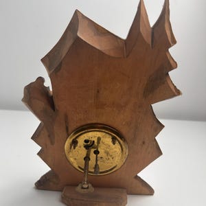 May include: A light brown wooden clock back featuring an irregular, jagged silhouette resembling a mountain or tree shape. A circular brass mechanism is mounted in the center, displaying two thin metal adjustment stems extending outward.