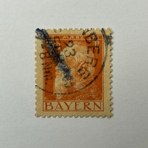 Könnte beinhalten: Eine orangefarbene Briefmarke mit einem Porträt und dem Wort "BAYERN" am unteren Rand. Die Briefmarke hat schwarze und blaue Markierungen und einen gezackten Rand. Der Text "3 MARZ" ist oben sichtbar.