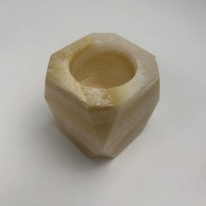 Op de afbeelding: Een zeshoekige kandelaar van lichtbeige steen. De kandelaar heeft een cirkelvormige opening aan de bovenkant en een facetgeslepen buitenkant. De steen heeft natuurlijke kleurvariaties, met strepen van donkerdere en lichtere tinten.