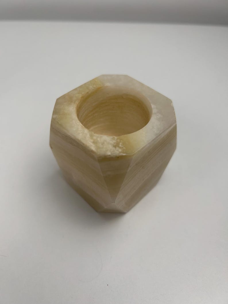 Op de afbeelding: Een zeshoekige kandelaar van lichtbeige steen. De kandelaar heeft een cirkelvormige opening aan de bovenkant en een facetgeslepen buitenkant. De steen heeft natuurlijke kleurvariaties, met strepen van donkerdere en lichtere tinten.