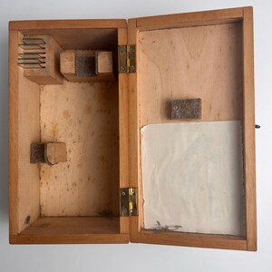 K&ouml;nnte beinhalten: Eine offene rechteckige Holzschachtel mit einem Klappdeckel, der zwei messingfarbene Metallscharniere aufweist. Das Innere enth&auml;lt ein kleines Holzgestell mit d&uuml;nnen Metallteilern, zwei filzgepolsterte St&uuml;tzen und eine wei&szlig;e Papiereinlage.