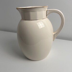 May include: A cream-coloured ceramic jug with a rounded body and a wide spout. The rim and the handle are accented with thin gold-coloured trim. The upper section of the jug features a subtle, faceted geometric design around the neck.
