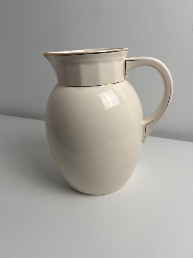 May include: A cream-coloured ceramic jug with a rounded body and a wide spout. The rim and the handle are accented with thin gold-coloured trim. The upper section of the jug features a subtle, faceted geometric design around the neck.