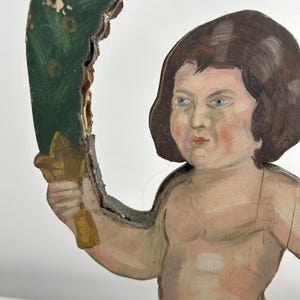 May include: A vintage, hand-painted wooden cherub figure with brown hair and blue eyes. The cherub holds a gold-colored object and is framed by a green, leaf-like arch. The paint shows signs of age and wear, adding to its antique charm.