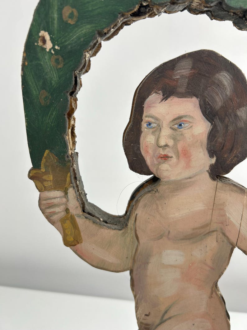 May include: A vintage, hand-painted wooden cherub figure with brown hair and blue eyes. The cherub holds a gold-colored object and is framed by a green, leaf-like arch. The paint shows signs of age and wear, adding to its antique charm.