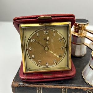 Puede incluir: Un reloj de viaje dorado con esfera cuadrada, alojado en un estuche de cuero rojo. La esfera del reloj tiene números y manecillas negras. El reloj descansa sobre un libro oscuro y envejecido.