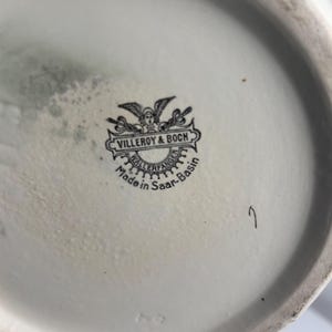May include: A white ceramic base featuring a black printed maker's mark for Villeroy & Boch. The stamp includes a winged figure, the text Villeroy & Boch, Wallerfangen, and Made in Saar-Basin. A small handwritten number 7 is visible nearby.