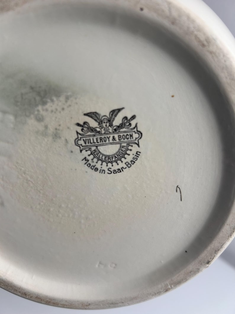 May include: A white ceramic base featuring a black printed maker's mark for Villeroy & Boch. The stamp includes a winged figure, the text Villeroy & Boch, Wallerfangen, and Made in Saar-Basin. A small handwritten number 7 is visible nearby.