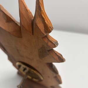 May include: A carved wooden object shaped like a stylized leaf or flame with a light brown finish. The piece features visible wood grain and textured edges. A circular brass-toned metal base is attached to the lower section with a small screw.