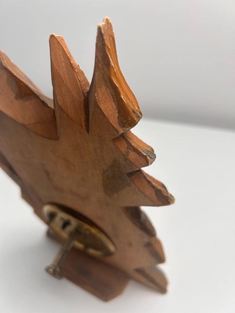 May include: A carved wooden object shaped like a stylized leaf or flame with a light brown finish. The piece features visible wood grain and textured edges. A circular brass-toned metal base is attached to the lower section with a small screw.