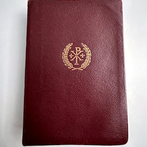 Könnte beinhalten: Ein rechteckiges Buch mit einem dunkelburgunderroten, strukturierten Einband. In der Mitte des Vorderdeckels befindet sich ein goldenes, geprägtes Emblem mit einem Chi-Rho-Symbol, das von einem Lorbeerkranz umgeben ist. Der Text Bavarian Heritage ist unten.