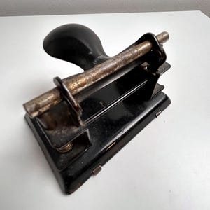 May include: A vintage black metal two-hole paper punch featuring a rounded handle and a weathered, oxidized metal rod across the top. The base is rectangular with a glossy black finish, showing signs of age and wear on the metallic components.