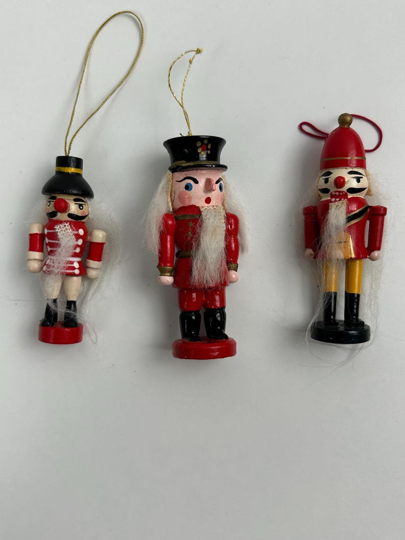 Könnte beinhalten: Drei hölzerne Weihnachts-Nussknacker-Ornamente. Jeder Nussknacker ist mit roten, schwarzen und weißen Details bemalt. Die Nussknacker haben goldene Schnuraufhänger. Die Nussknacker stehen auf einer weißen Oberfläche.