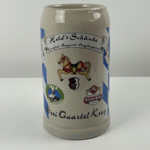 May include: A white ceramic beer stein with a blue and white checkered pattern. The stein features a colorful illustration of a carousel horse, a logo for "Held's Schänke", and the text "Königlich-Bayrisch-Augsburgerisch". The stein also has logos for "IHLE", "Hasen-Brau", and the text "Brei Quartel Krug".