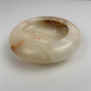 Op de afbeelding: Een ronde, gepolijste asbak van cr&egrave;mewitte onyx met subtiele oranje en bruine adering. De asbak heeft een gladde, glanzende afwerking en een ondiepe, cirkelvormige kom. Het ontwerp is eenvoudig en elegant.