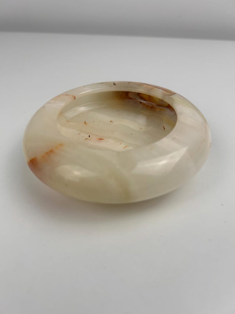Op de afbeelding: Een ronde, gepolijste asbak van cr&egrave;mewitte onyx met subtiele oranje en bruine adering. De asbak heeft een gladde, glanzende afwerking en een ondiepe, cirkelvormige kom. Het ontwerp is eenvoudig en elegant.