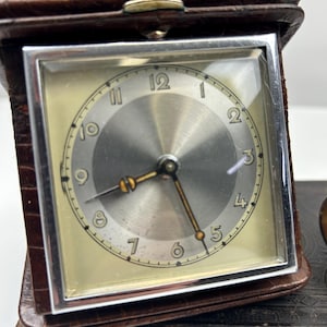 Puede incluir: Un reloj de viaje vintage con una esfera cuadrada plateada y manecillas doradas, en un estuche de cuero marrón. La esfera del reloj presenta números negros y un segundero. El estuche está grabado con un patrón de cocodrilo.