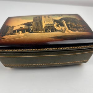 以下が含まれることがあります： 光沢のある濃い茶色の蓋に、建物と記念碑のセピア色の画像が描かれた長方形の小物入れ。ボックスは、金色のトリムと小さな足が付いた濃い緑色のベースです。 全体的な美学はヴィンテージです。