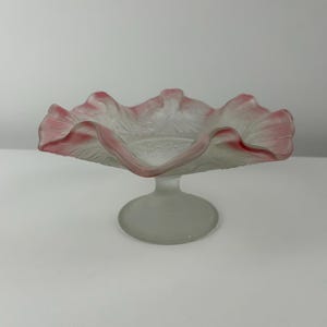 Puede incluir: Un cuenco de pedestal de vidrio esmerilado con un borde rizado y un borde rosa. El cuenco está sobre un fondo blanco.