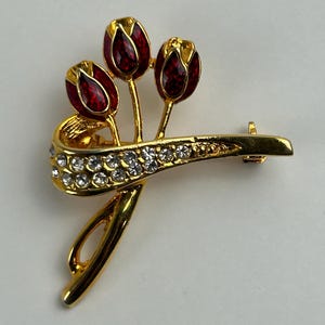 May include: A gold-toned brooch featuring three red and gold tulip-shaped flowers. The brooch has a curved design with clear crystal accents. The pin is a decorative accessory.
