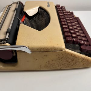 Może przedstawiać: Beżowa przenośna maszyna do pisania z nakrapianym wykończeniem i ciemnobordowymi klawiszami. Urządzenie posiada układ klawiatury QWERTY, czarny wałek, metalową podp&oacute;rkę na papier oraz chromowaną dźwignię powrotu karetki po lewej stronie.