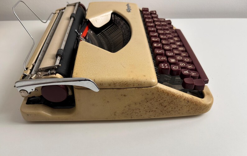Może przedstawiać: Beżowa przenośna maszyna do pisania z nakrapianym wykończeniem i ciemnobordowymi klawiszami. Urządzenie posiada układ klawiatury QWERTY, czarny wałek, metalową podp&oacute;rkę na papier oraz chromowaną dźwignię powrotu karetki po lewej stronie.