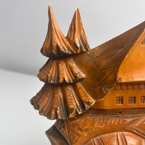 May include: A carved wooden clock featuring a chalet-style house design with a pointed roof and a stylized evergreen tree attached to the side. The wood has a warm brown finish with darker accents in the crevices of the intricate carvings.