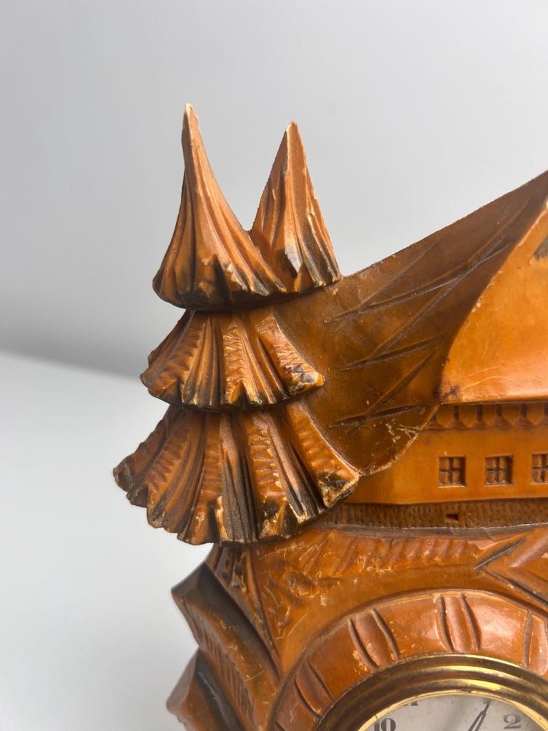 May include: A carved wooden clock featuring a chalet-style house design with a pointed roof and a stylized evergreen tree attached to the side. The wood has a warm brown finish with darker accents in the crevices of the intricate carvings.