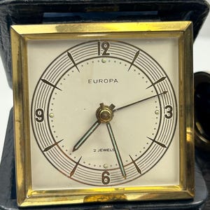 Puede incluir: Un reloj de viaje Europa vintage con un marco cuadrado dorado y una esfera color crema. El reloj presenta números negros, manecillas y el texto "EUROPA" y "2 JEWELS". Está encerrado en un estuche de viaje de polipiel negro.