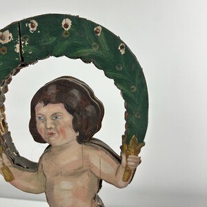 May include: A vintage wooden cherub figurine with a green and gold halo. The cherub has brown hair, blue eyes, and is holding the halo with both hands. The halo is painted green with white accents.