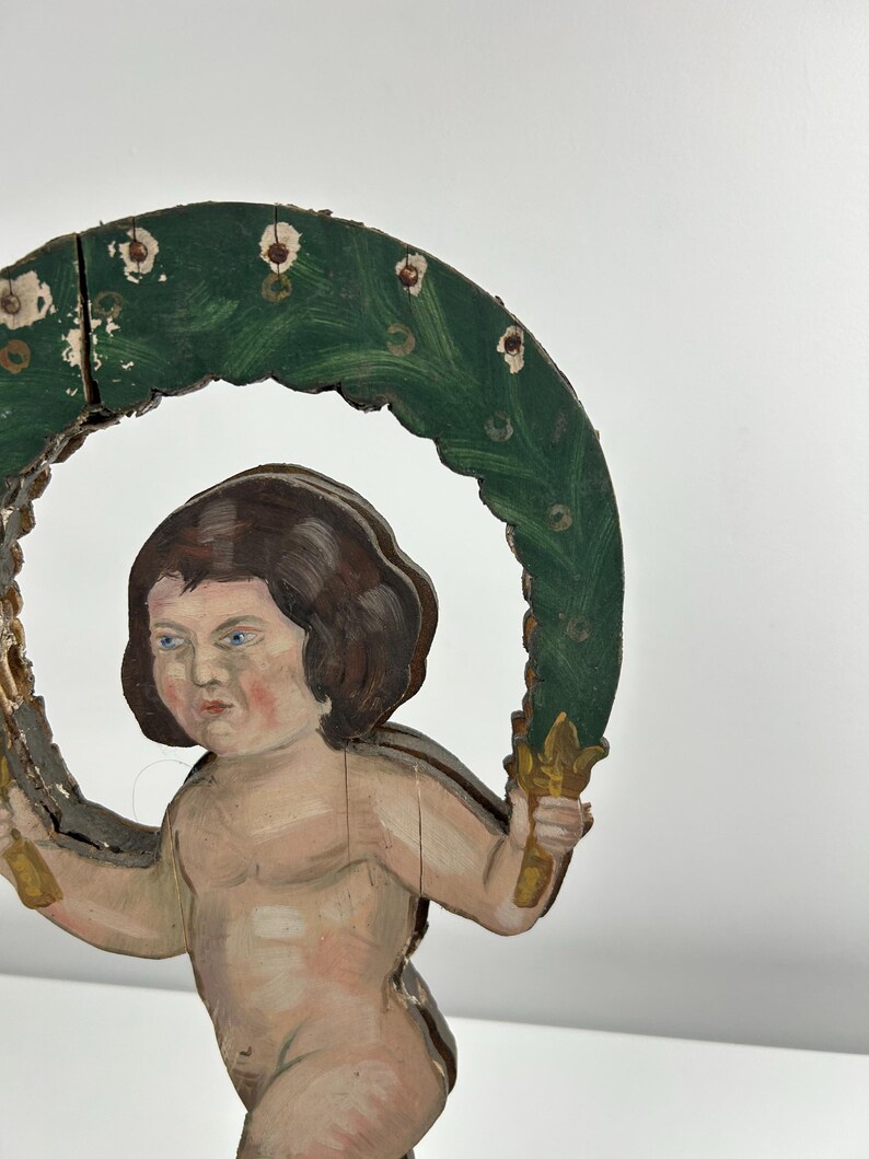 May include: A vintage wooden cherub figurine with a green and gold halo. The cherub has brown hair, blue eyes, and is holding the halo with both hands. The halo is painted green with white accents.