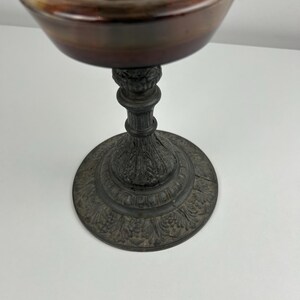May include: An antique oil lamp with a silver-colored top and a dark brown base. The lamp has a decorative, ornate base with a detailed floral pattern. The lamp is made of metal and sits on a white surface.