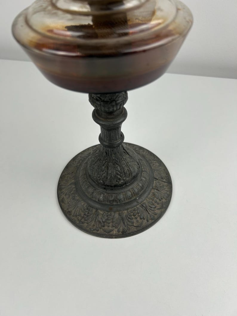 May include: An antique oil lamp with a silver-colored top and a dark brown base. The lamp has a decorative, ornate base with a detailed floral pattern. The lamp is made of metal and sits on a white surface.