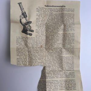 K&ouml;nnte beinhalten: Eine alte, cremefarbene Bedienungsanleitung aus Papier mit einer schwarz-wei&szlig;en Strichzeichnung eines Mikroskops auf der linken Seite. Das Dokument enth&auml;lt umfangreichen deutschen Text mit dem Titel Gebrauchsanweisung und endet mit Made in Germany.