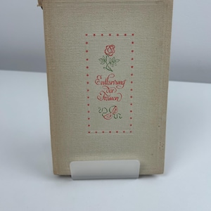 May include: A vintage book with a textured, cream-coloured cover. The cover features a red and green floral design with the German text "Entlarvung der Frauen" in a decorative font, surrounded by red star accents. The book is propped up on a clear stand.