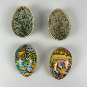 Op de afbeelding: Vier vintage papieren paaseieren dozen. Twee dozen zijn open en laten een groen bloemmotief zien aan de binnenkant. De andere twee dozen zijn gesloten en laten kleurrijke illustraties van dieren en scènes zien. Eén doos laat een haan, een kikker en eenden zien. De andere doos laat een konijn en kuikens in een keuken zien.