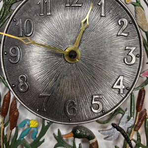 Puede incluir: Una esfera de reloj plateada con números romanos negros y marcadores de minutos negros. La esfera del reloj está rodeada por un marco de metal con un diseño floral verde, marrón y azul. La esfera del reloj está situada sobre un fondo de dos patos, una libélula y juncos.