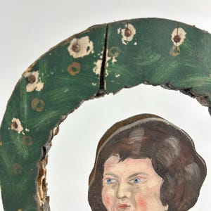 May include: A vintage, hand-painted wooden ornament featuring a cherubic figure with blue eyes and brown hair. The figure is partially framed by a green, wreath-like element with white and gold accents. The wood shows signs of age and wear.