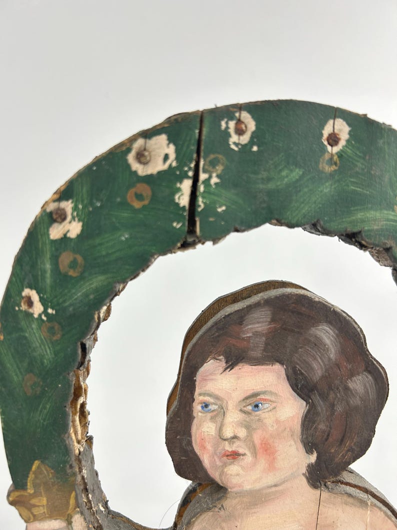 May include: A vintage, hand-painted wooden ornament featuring a cherubic figure with blue eyes and brown hair. The figure is partially framed by a green, wreath-like element with white and gold accents. The wood shows signs of age and wear.