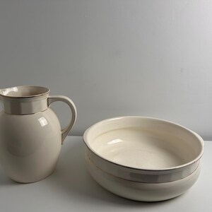 May include: A cream-coloured ceramic pitcher with a rounded body and a matching wide, shallow ceramic basin. Both pieces feature a subtle, thin metallic gold rim along the top edges, providing a clean and uniform appearance for this set.