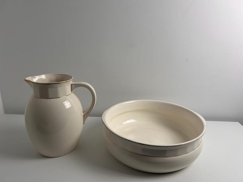 May include: A cream-coloured ceramic pitcher with a rounded body and a matching wide, shallow ceramic basin. Both pieces feature a subtle, thin metallic gold rim along the top edges, providing a clean and uniform appearance for this set.