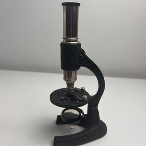 K&ouml;nnte beinhalten: Ein Mikroskop im Vintage-Stil mit einem strukturierten, schwarzen Gussmetallrahmen, einem silberfarbenen zylindrischen Okular, einem runden Objekttisch mit zwei Metallklammern und einem kleinen, verstellbaren runden Spiegel am Fu&szlig;.