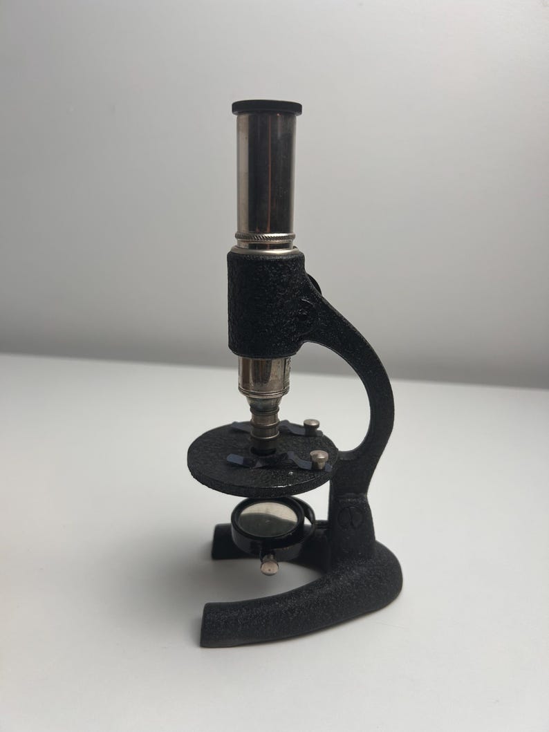 K&ouml;nnte beinhalten: Ein Mikroskop im Vintage-Stil mit einem strukturierten, schwarzen Gussmetallrahmen, einem silberfarbenen zylindrischen Okular, einem runden Objekttisch mit zwei Metallklammern und einem kleinen, verstellbaren runden Spiegel am Fu&szlig;.